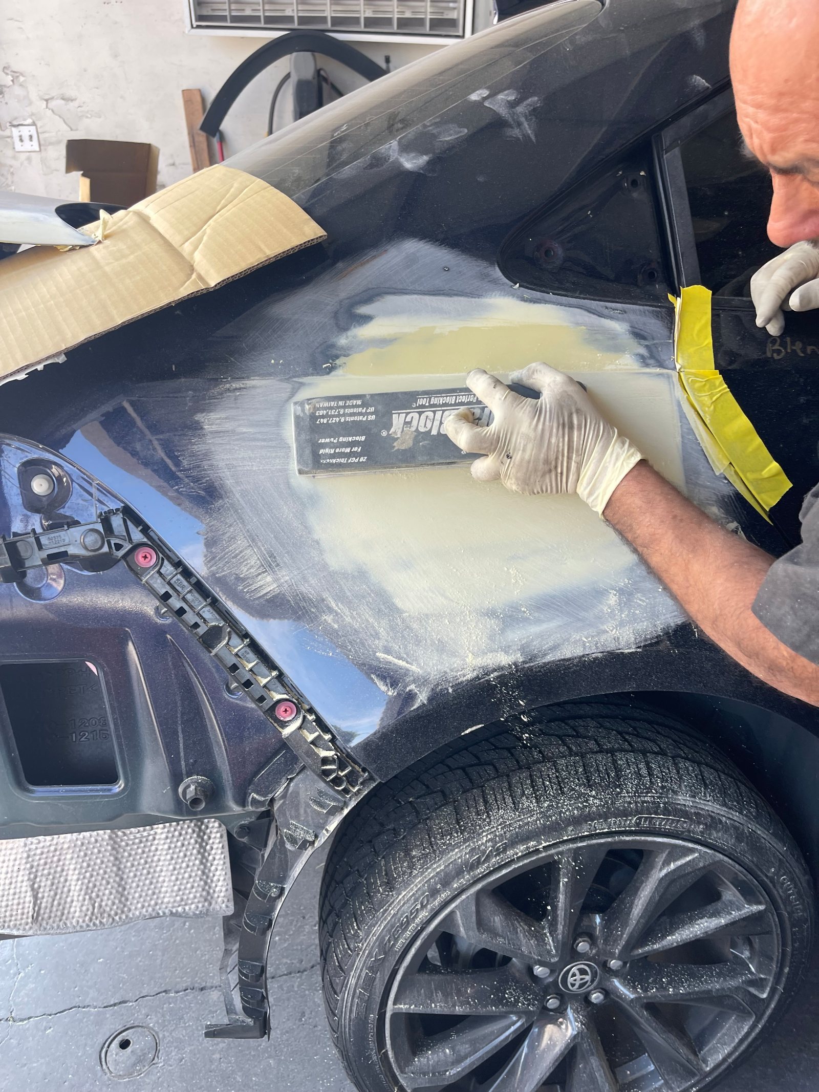 Close crop on the gloved hands blocking filler on that same navy quarter panel with a Dura-Block, tool and hands filling
