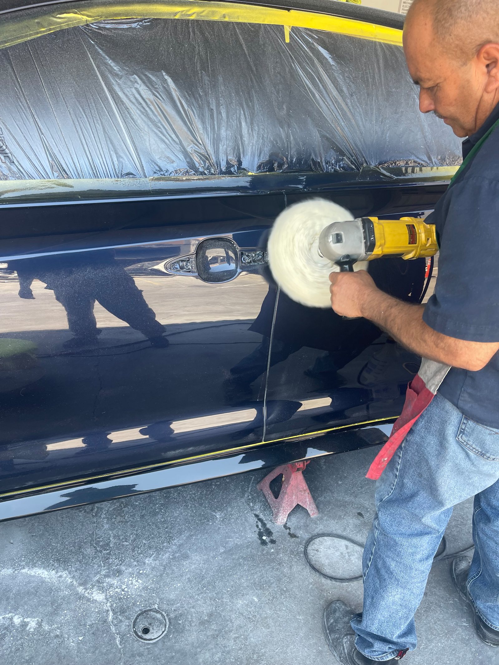 The technician buffing the freshly refinished flank of the dark navy sedan with a rotary polisher and wool pad, glass masked